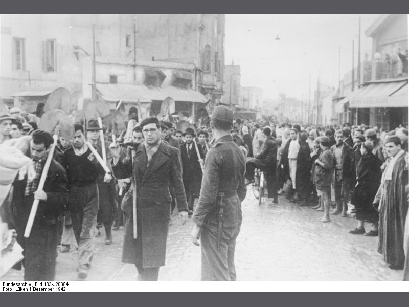 Kolonne mit jüdischen Zwangsarbeitern unter den Augen der muslimischen Bevölkerung in Tunesien im Dezember 1942. (Quelle: Bundesarchiv, Bild 183-J20384 / CC-BY-SA 3.0 /WikiCommons)